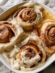 Extreme close-up of jumbo cinnamon rolls submerged in golden-brown heavy cream sauce with thick cream cheese icing. (cinnamon roll breakfast bake)
