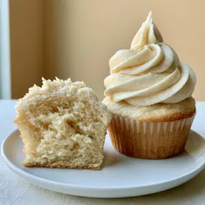 A close up of a vanilla cupcake sliced open revealing a light yellow moist crumb interior on a white plate. (Vanilla Cupcakes)