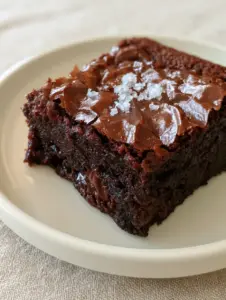 An unedited handheld smartphone photo of a thick, ultra-fudgy chocolate brownie on a clean cream ceramic plate showing a delicate, paper-thin, shiny crinkle-top crust with tiny fracture lines. (brownie recipe)